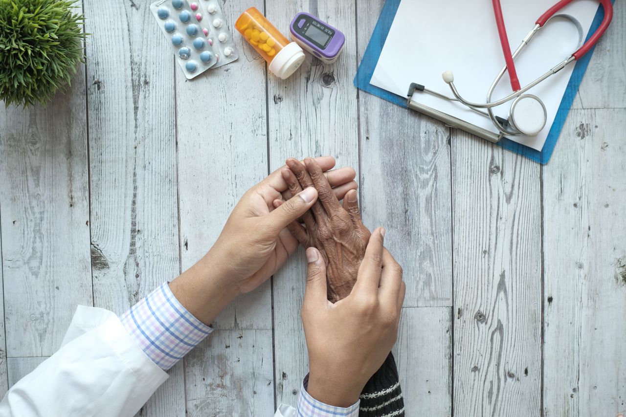 A doctor holding an elderly patients hands on a desk with medication and stethoscope.